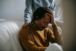Upset man with his hand on his head, being comforted by someone sitting bhind him. 
